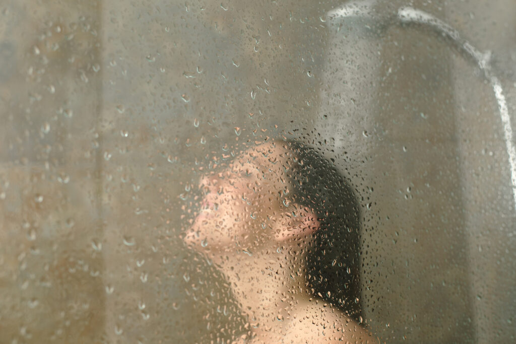 Woman having shower behind glass with water drops standing on a bathroom; 