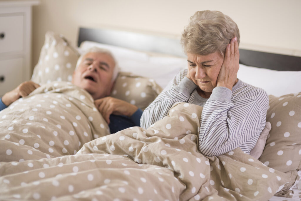Older couple in bed. Woman has hands over her ears, man is lying on his back snoring