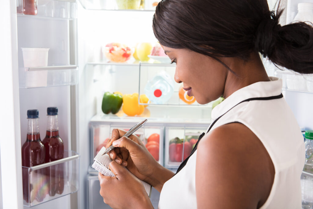 Close-up Of Young African Woman Writing On Spiral Book Near Open Fridge