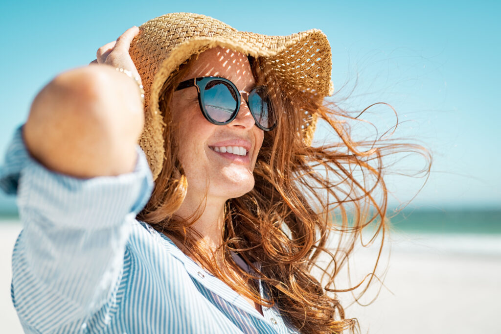 Side view of beautiful mature woman wearing sunglasses at beach. Young smiling woman on vacation looking away while enjoying sea breeze wearing straw hat. Closeup portrait of attractive girl relaxing.; 