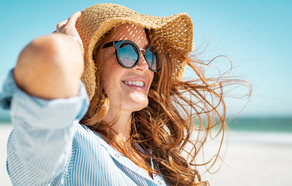 Side view of beautiful mature woman wearing sunglasses at beach. Young smiling woman on vacation looking away while enjoying sea breeze wearing straw hat. Closeup portrait of attractive girl relaxing.;