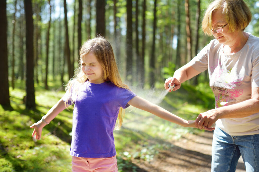 Middle age woman applying insect repellent to her granddaughter before forest hike beautiful summer day. Protecting children from biting insects at summer. Active leisure with kids.; 