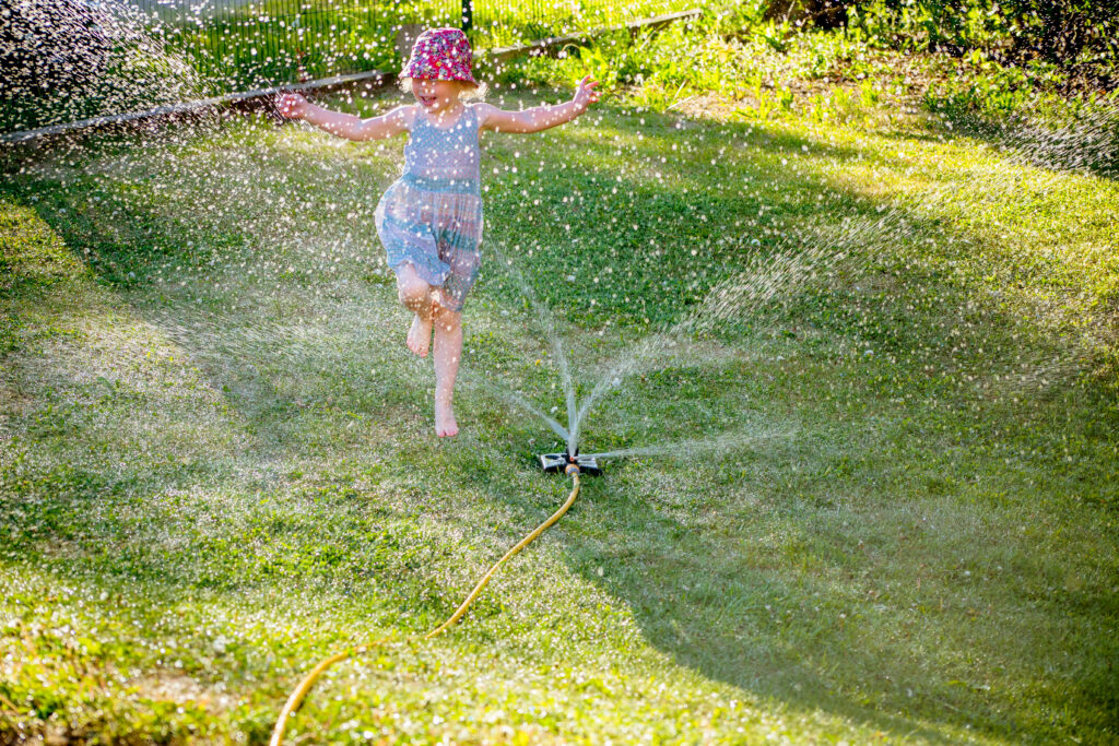 Irrigation sprinkler device for irrigation of home lawn, grass working and happy playful girl jumps around and plays with splashing water in summer.; 