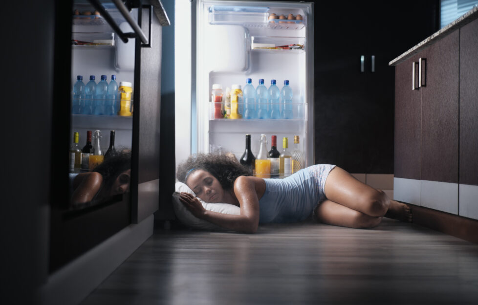 Young hispanic woman suffering for summer heat and lack of air conditioning at home. Black girl covered with sweat sleeping on floor with head inside fridge.;