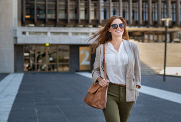 Portrait of successful happy woman on her way to work on street. Confident business woman wearing blazer carrying side bag walking with a smile. Smiling woman wearing sunglasses and walking on street.;