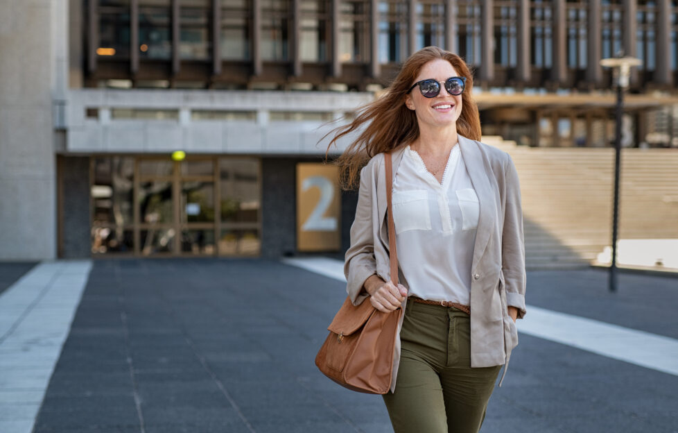 Portrait of successful happy woman on her way to work on street. Confident business woman wearing blazer carrying side bag walking with a smile. Smiling woman wearing sunglasses and walking on street.;