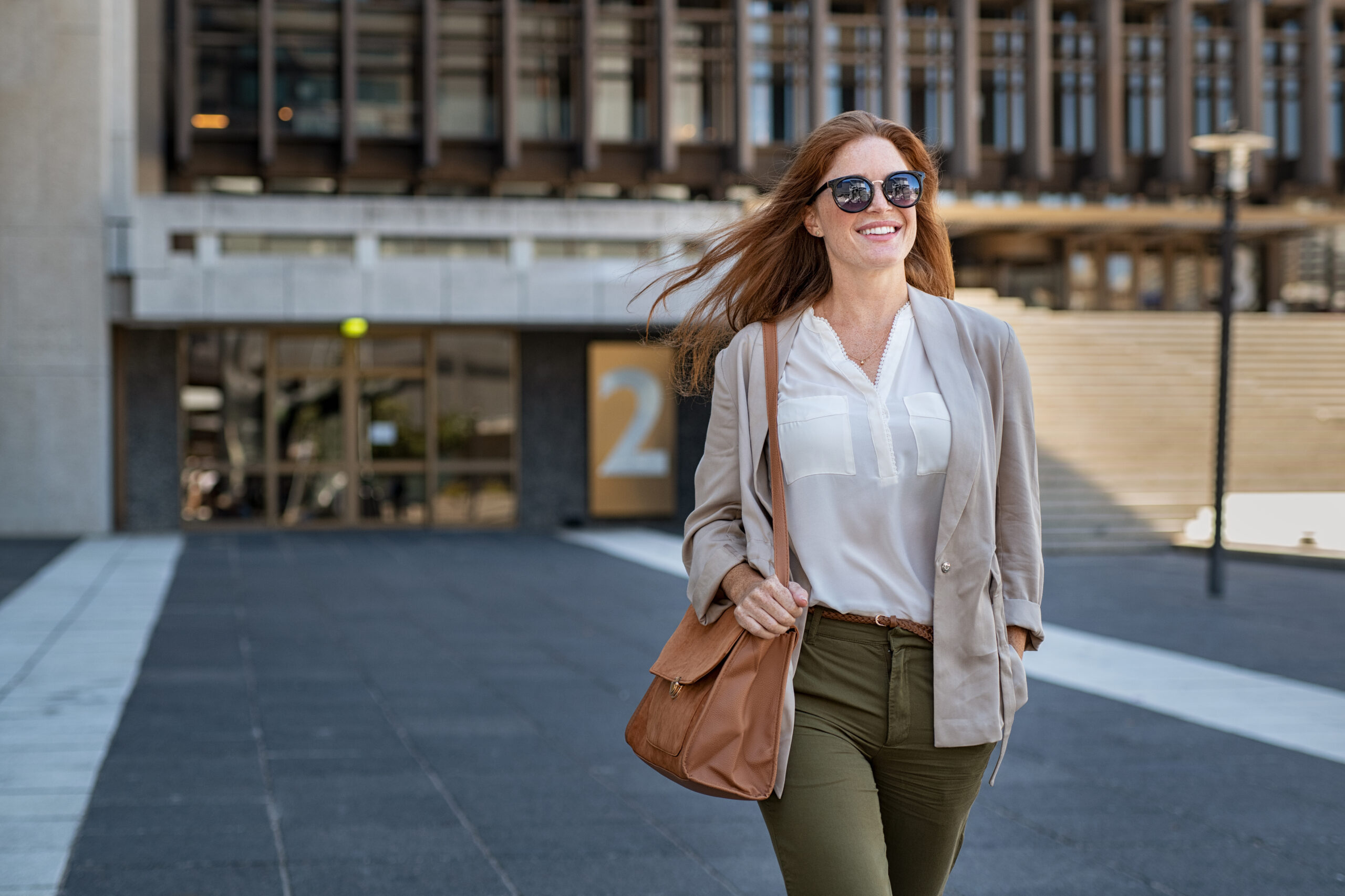 Portrait of successful happy woman on her way to work on street. Confident business woman wearing blazer carrying side bag walking with a smile. Smiling woman wearing sunglasses and walking on street.;