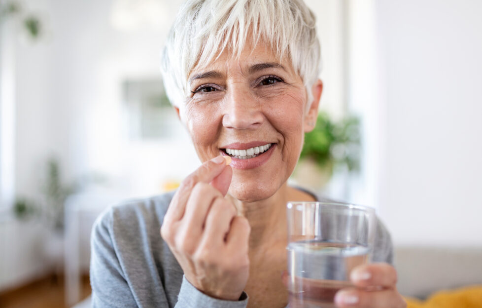 Head shot portrait happy woman holds pill glass of water, takes daily medicine vitamin D, omega 3 supplements, skin hair nail strengthen and beauty, medication for health care concept;