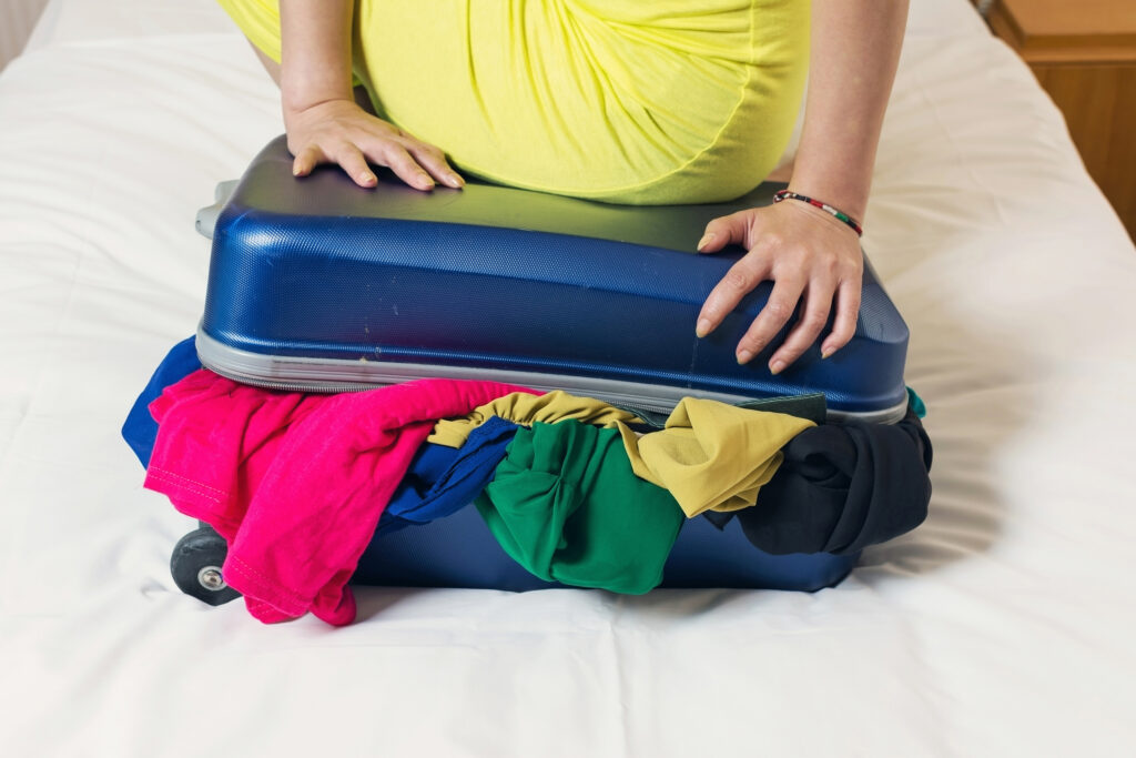 Young woman try to close the overfilled suitcase in the room.;