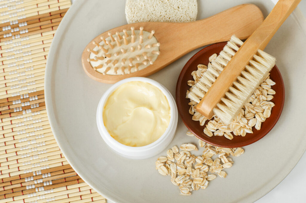 Yellow hair mask (banana face cream, shea butter facial mask, body butter) in the small white jar, oatmeal, massage body brush, wooden hairbrush. Natural skin and hair concept. Top view, copy space.; 