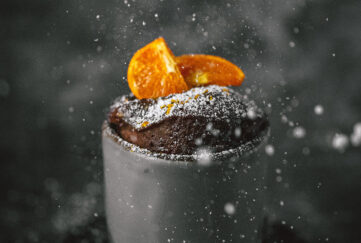 A fluffy chocolate cake with orange segments on top can be seen rising out of a grey mug. There's a plain black background with swirls of steam and icing sugar sprinkling down on top.