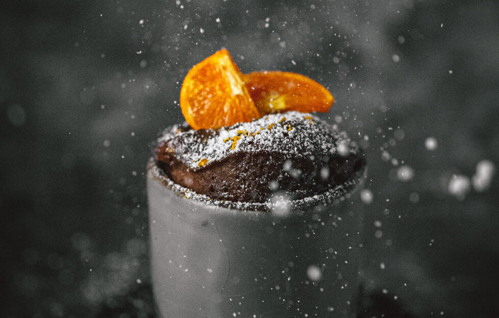 A fluffy chocolate cake with orange segments on top can be seen rising out of a grey mug. There's a plain black background with swirls of steam and icing sugar sprinkling down on top.