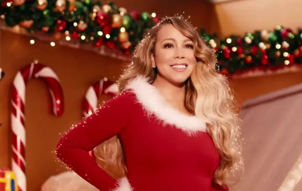 Image capture from Mariah Carey's updated music video for All I Want For Christmas, 2019. Features Mariah in front of a Christmas backdrop wearing a Santa suit and surrounded by sparkles. The song is a Christmas Number One Contender for 2023.