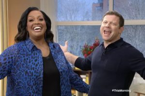 Image show Alison Hammond and Dermot O'Leary on the Friday show. They will continue to work alongside the new This Morning Presenters. Both have arms spread wide and smiling as they announce something with a lot of joy. Alison is wearing a blue leopard print shirt with a black top underneath. Dermot is wearing a plain navy button collared top.