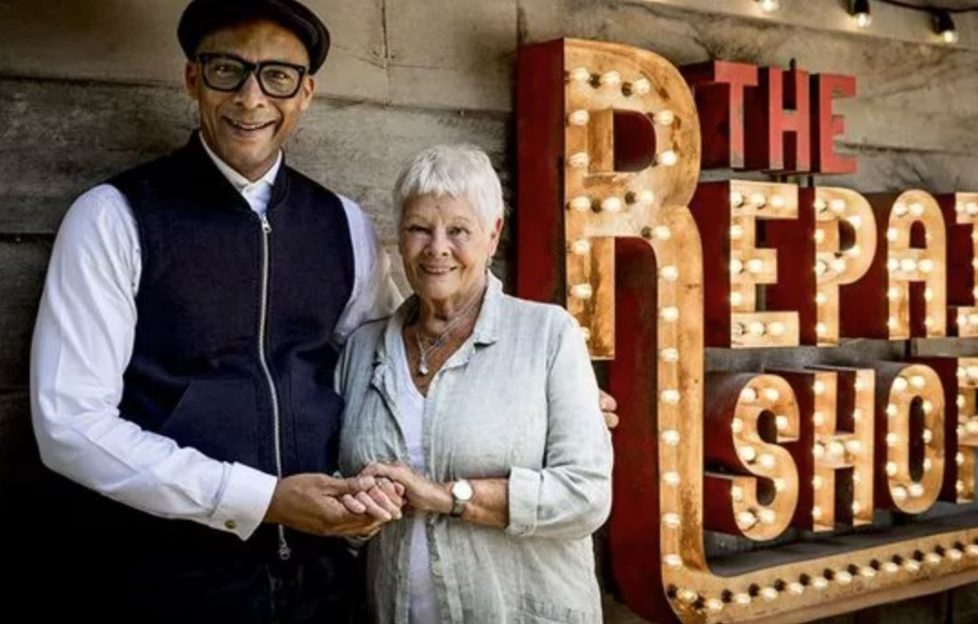 Image shows Judy Dench and Jay Blades standing in front of The Repair Shop illuminated sign. Jay his holding Judy in a warm embraces with her hand in his as the pair smile for the camera.