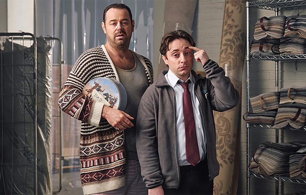 Image show promo shot for Sky's new comedy series Mr Bigstuff. The show stars Danny Dyer and Ryan Sampson who are seen here in character. Danny dyer's character is beaten up from a fight and wearing a knitted cardigan while holding a biscuit tin. Ryan's cahracter looks stressed in a shirt and tie and light grey jacket. New tv shows July.