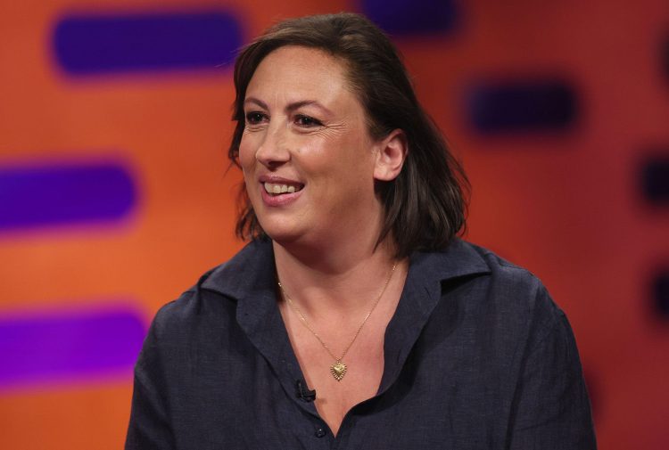 Miranda Hart discusses Lyme disease on Graham Norton talk show. She's wearing a black blouse and a silver necklace. the background is orange with purple oblongs.