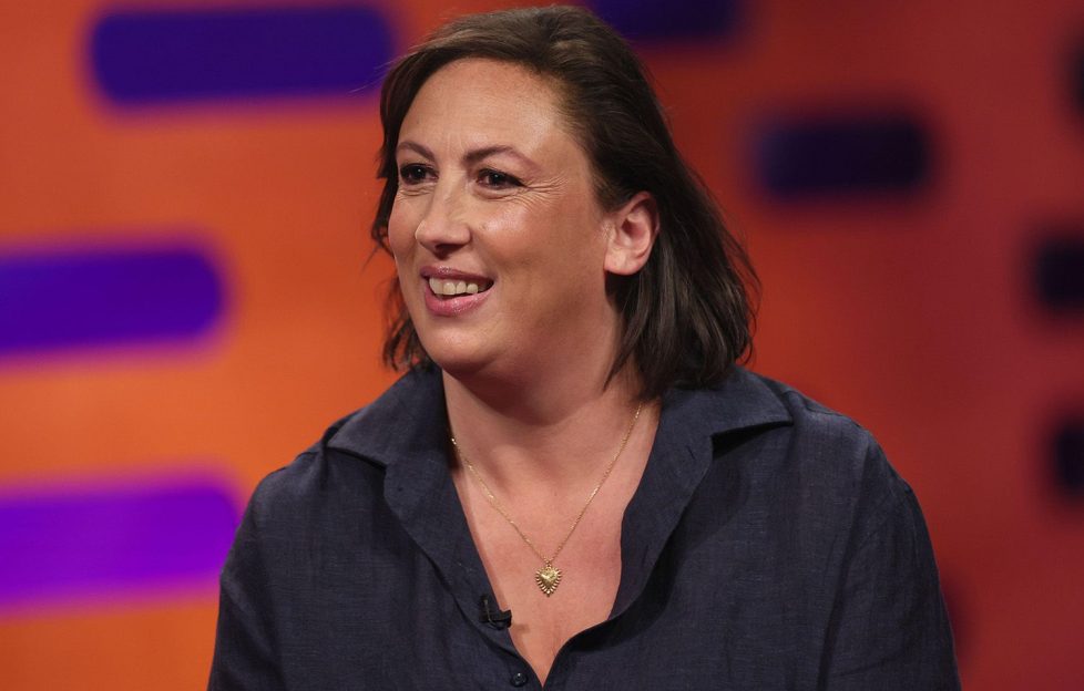 Miranda Hart discusses Lyme disease on Graham Norton talk show. She's wearing a black blouse and a silver necklace. the background is orange with purple oblongs.