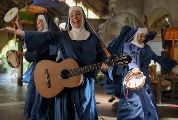 Still frame from Paddington in Peru starring Olivia Colman as a guitar playing nun. She is dancing and singing with other nuns all wearing blue robes.