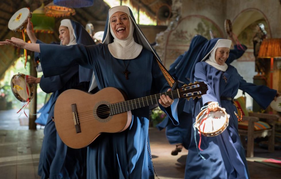 Still frame from Paddington in Peru starring Olivia Colman as a guitar playing nun. She is dancing and singing with other nuns all wearing blue robes.